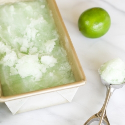 Homemade lime sorbet in a gold loaf pan. A lime and an ice cream scooper are nearby.