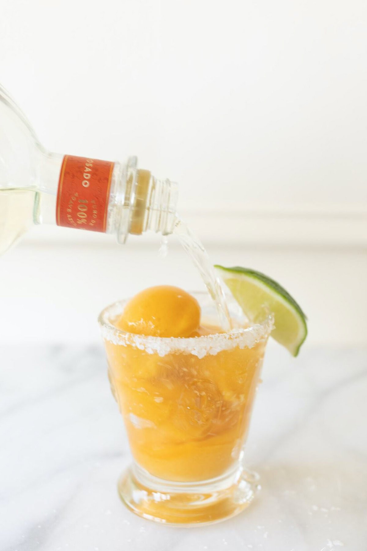 A glass with an orange drink and ice on a marble surface is garnished with a lime wedge. Clear liquid is being poured into the glass from a bottle, creating a refreshing margarita float.