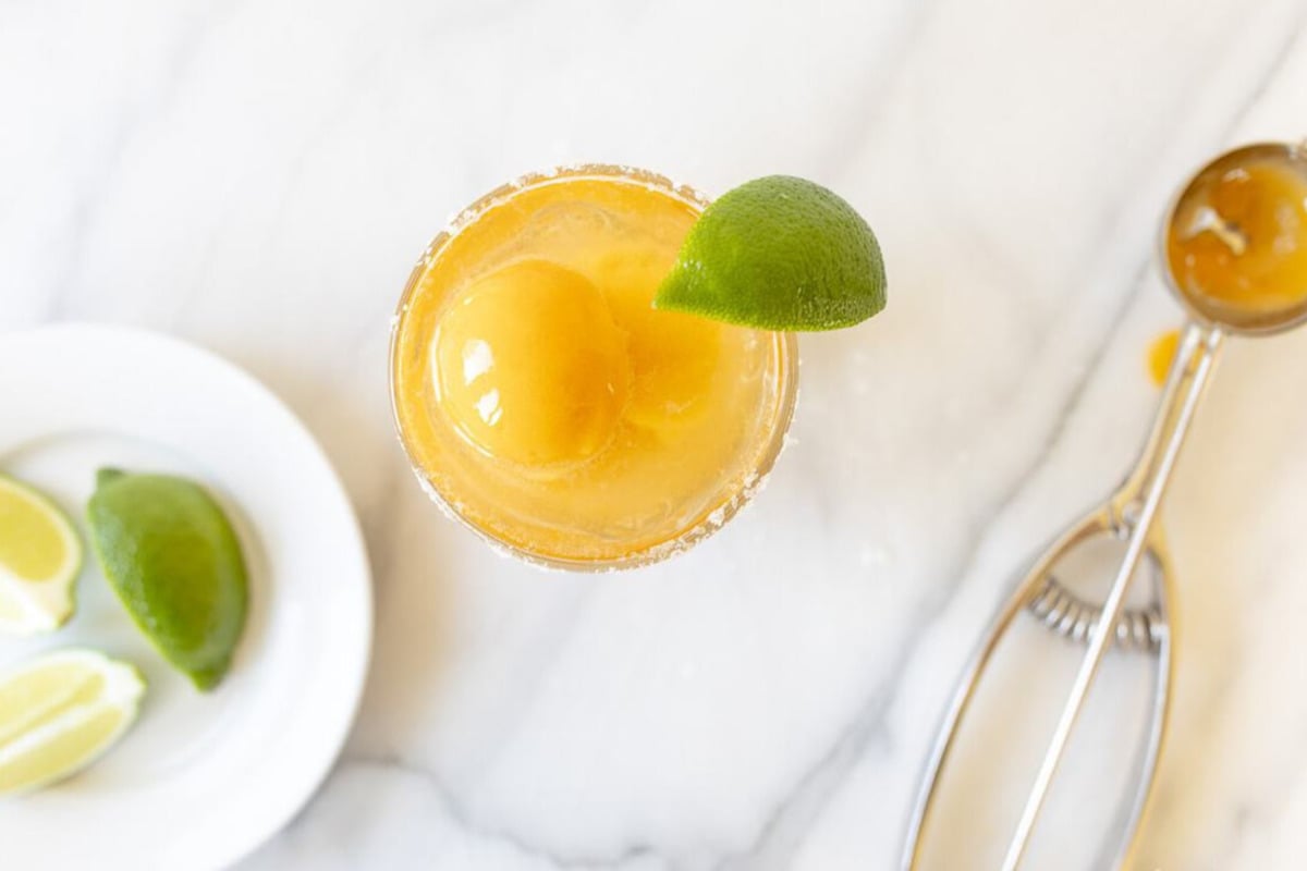 Top-down view of a margarita float with a lime wedge on the rim, garnished with a scoop of mango sorbet. A plate with lime wedges and a scooper with sorbet are placed beside the drink on a marble surface.