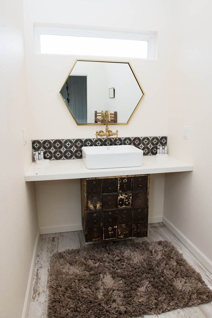 A modern industrial farmhouse bathroom with a raised sink and geometric brass mirror.