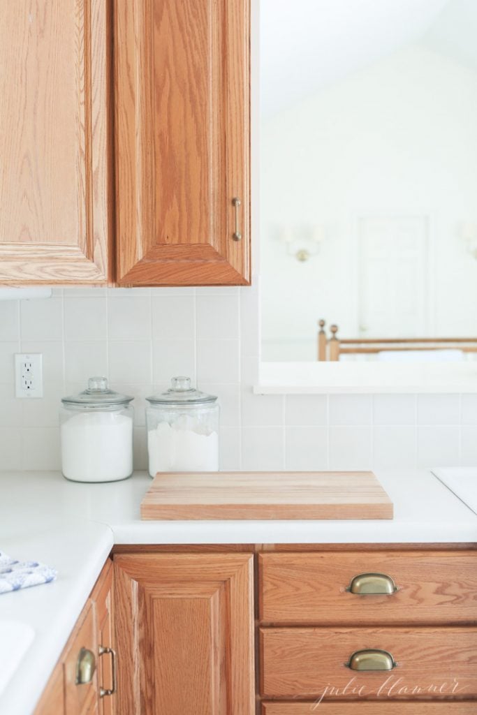 Brass hardware looks rich and warm against oak cabinets. Light-colored backsplash and counters with glass canisters of flour and sugar. 