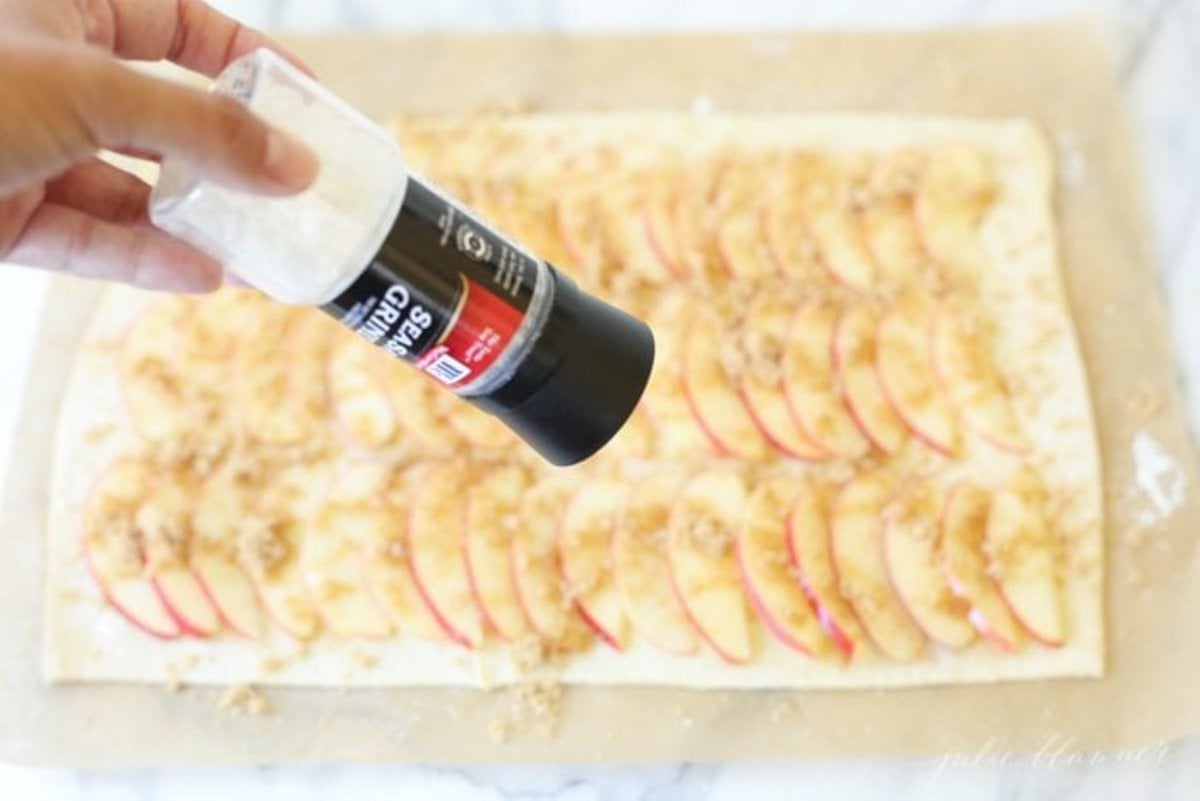 A hand grinding salt over a caramel apple tart before going into the oven