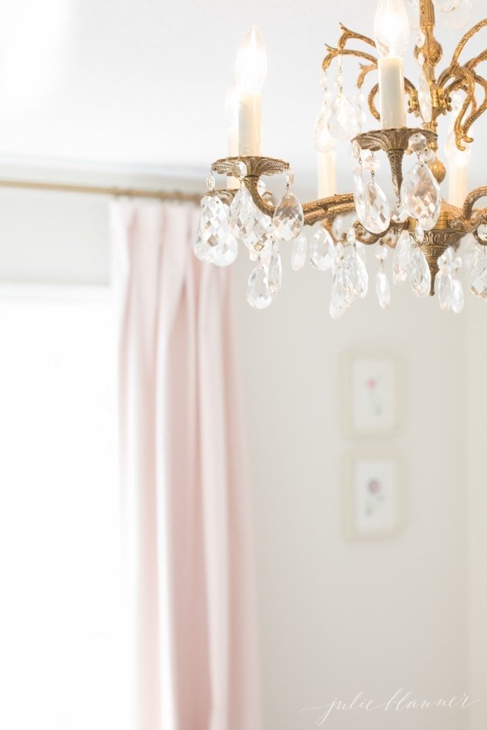 Pale pink curtains in the background of a white bedroom, vintage gold chandelier in focus of image.