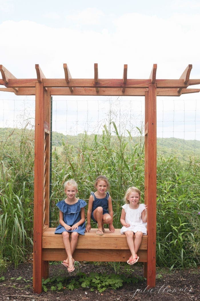 Three little girls sitting on a garden bench with grasses behind them.