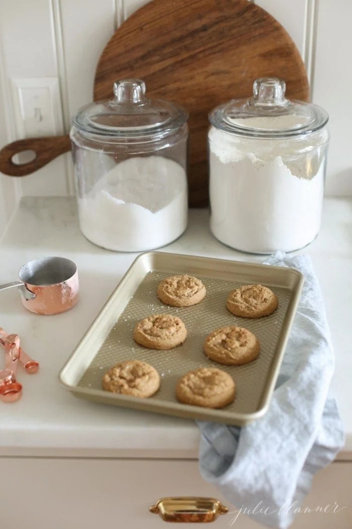 Brown sugar cookies on a gold baking sheet after coming out of the oven, flour and sugar canisters in background