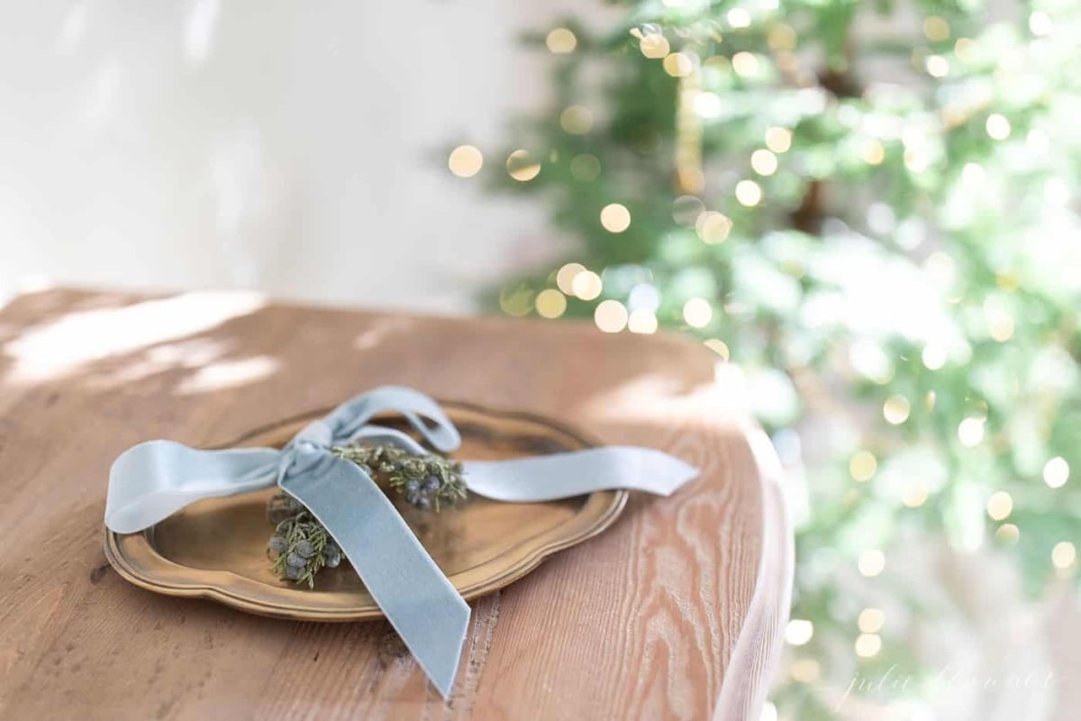 A gold plate with a bow on it sits on a table in front of a Christmas tree adorned with Christmas lights.