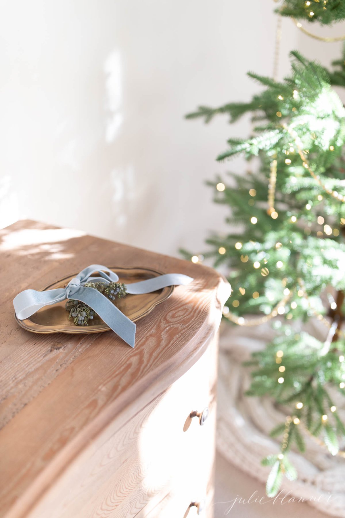 A wooden dresser with a blue ribbon on it next to a Christmas tree adorned with Christmas lights in a bedroom.