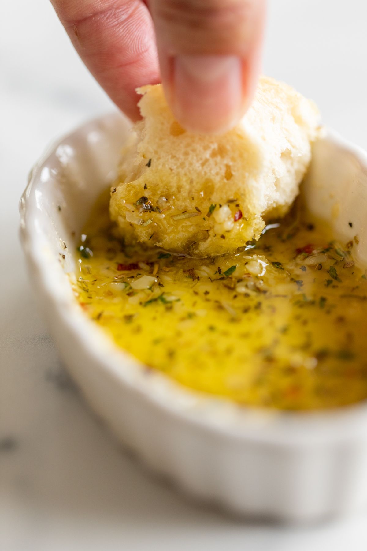 A hand dipping a piece of bread into a white ramekin full of olive oil bread dip.