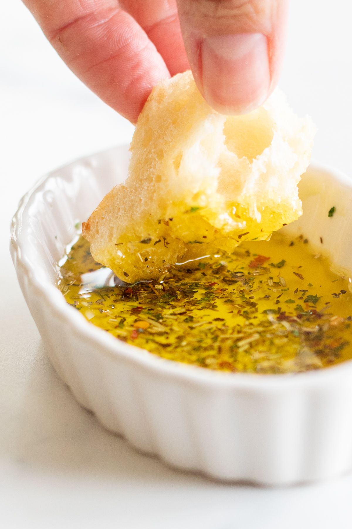 A hand dipping a piece of bread into a white ramekin full of olive oil bread dip.