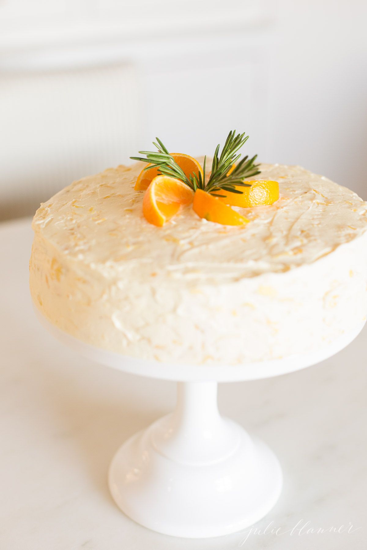 Sliced mandarin oranges and rosemary sprigs on top of a mandarin orange cake on a cake stand. 