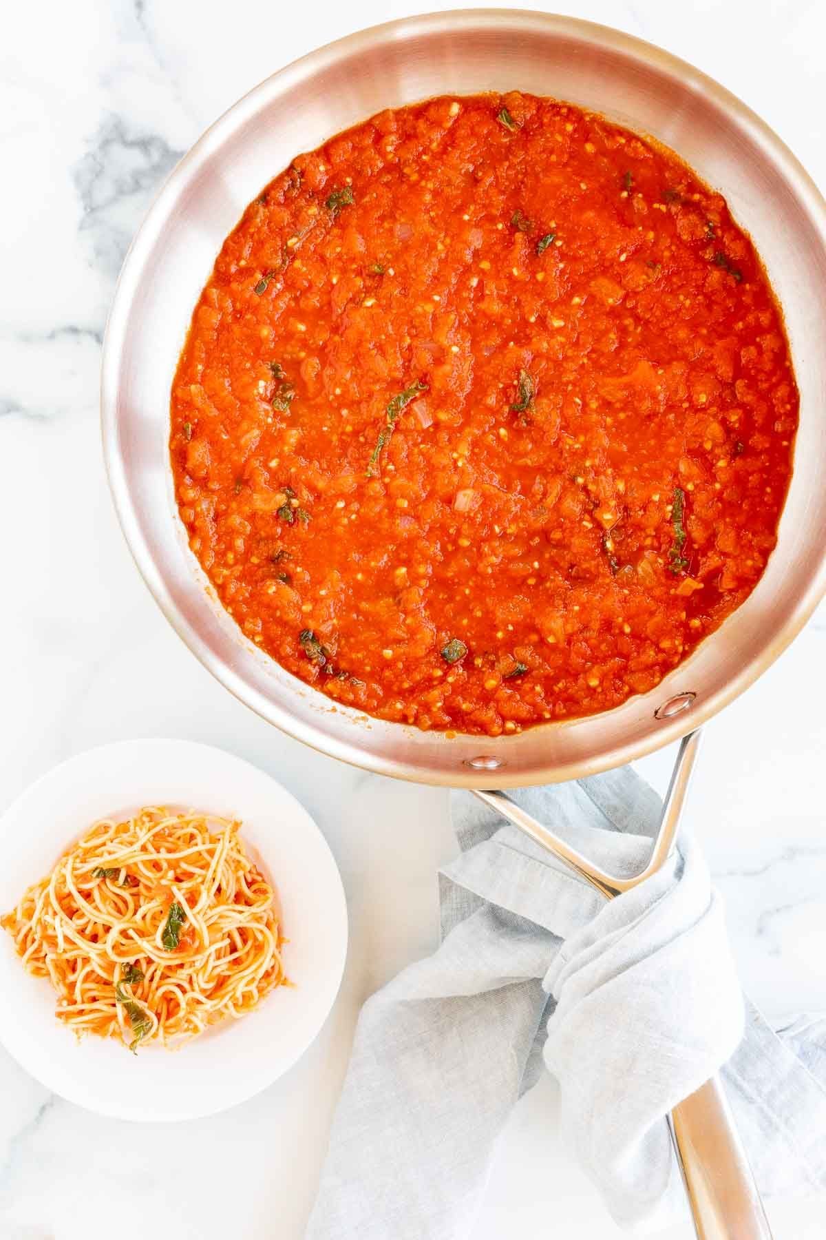 A silver saucepan full of pomodoro sauce, a plate of pomodoro pasta to the side