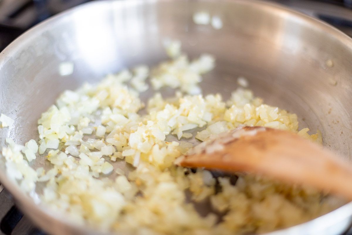 Chopped onions cooking in a silver pan with a wooden spoon.