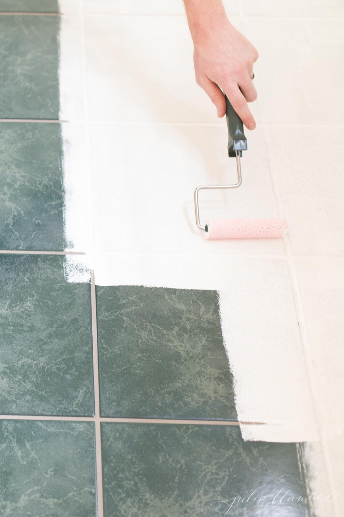 A hand, using a paint roller to roll white tile paint over old green tiles. 