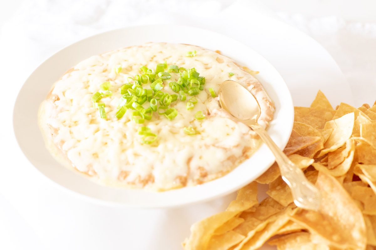 A white bowl full of cream cheese bean dip, topped with diced chives and tortilla chips to the side