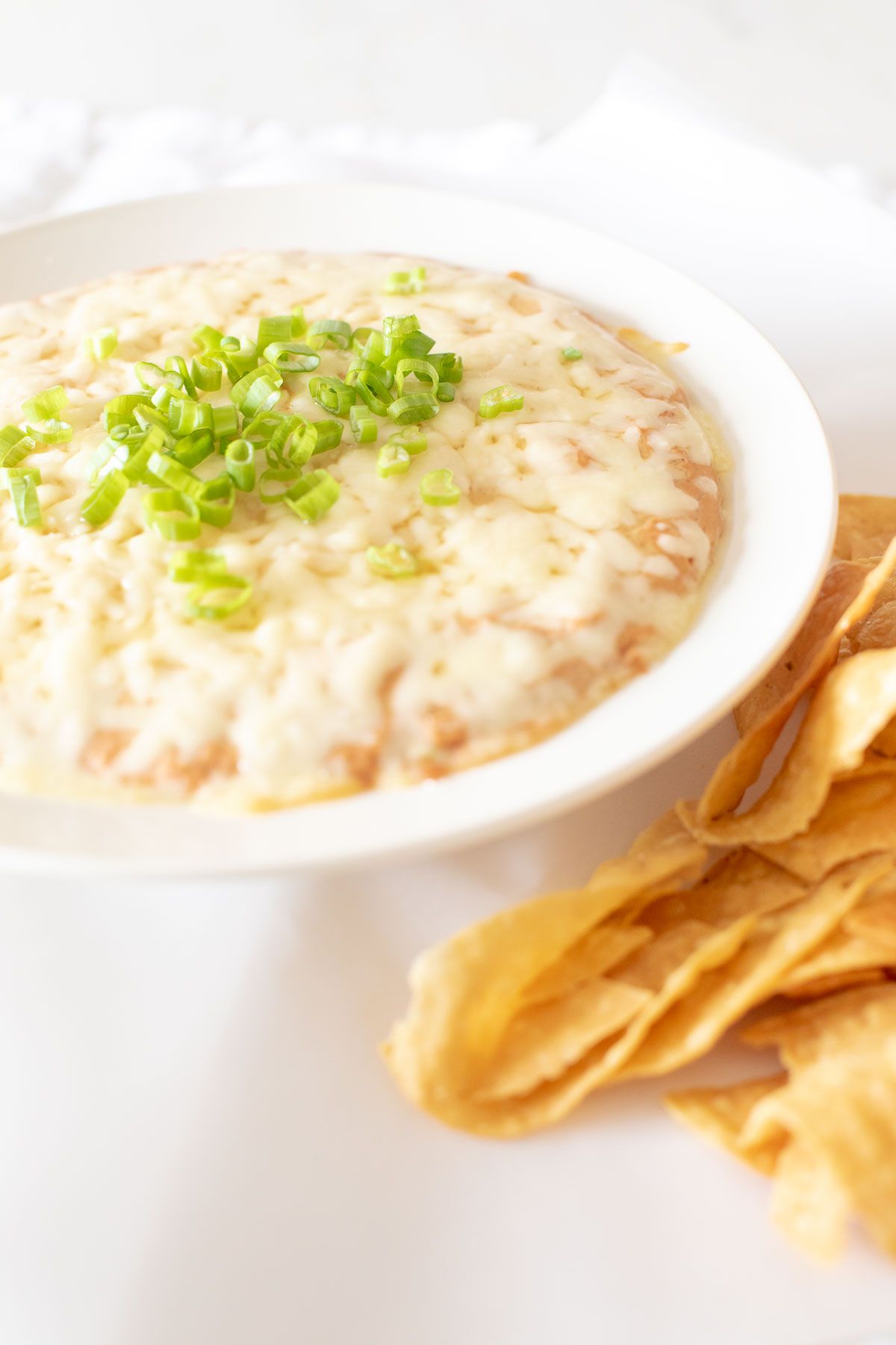 A white bowl full of cream cheese bean dip, topped with diced chives and tortilla chips to the side