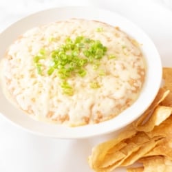 A white bowl full of cream cheese bean dip, topped with diced chives and tortilla chips to the side