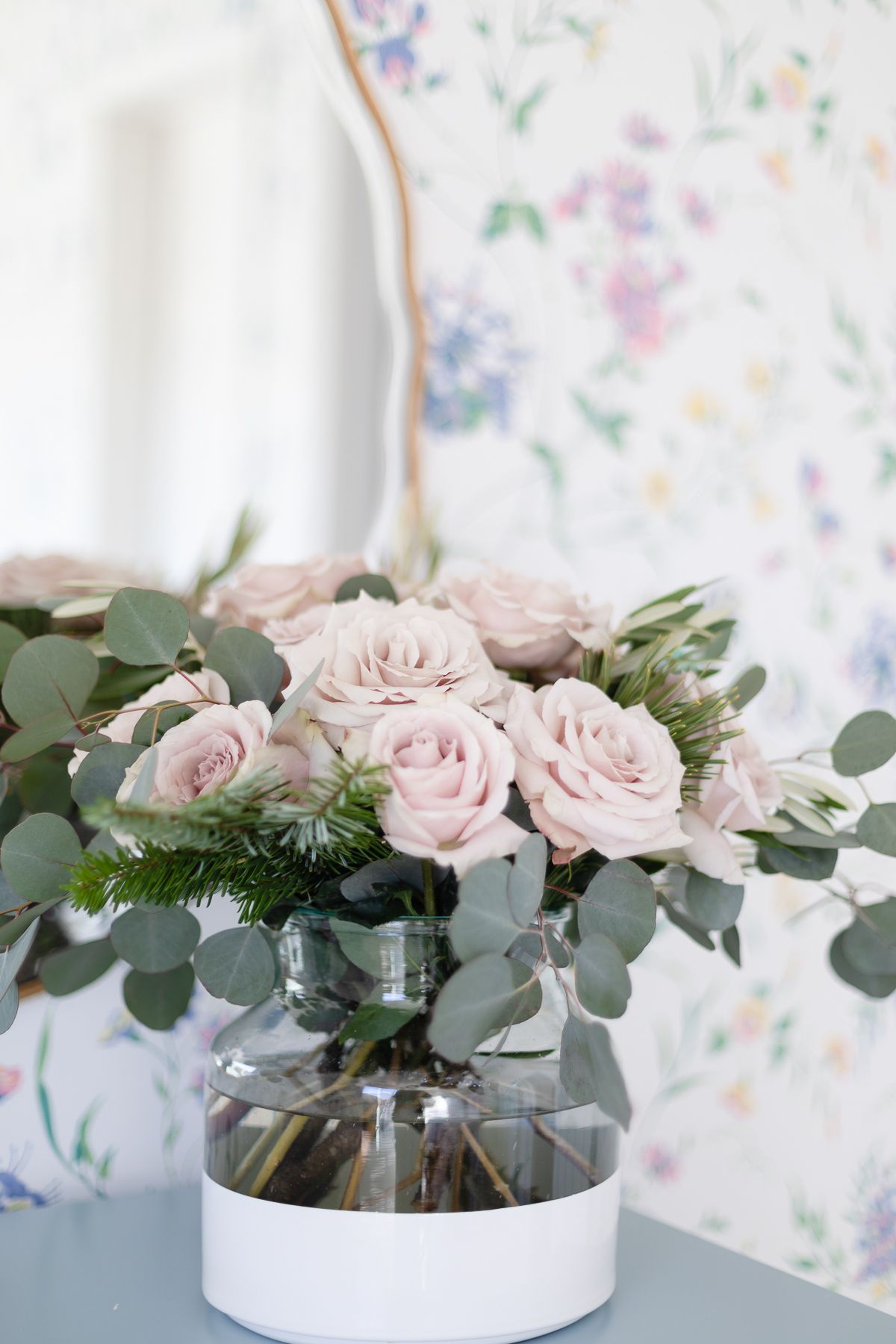 Pink roses in a white dipped flower vase in a wallpapered bedroom.