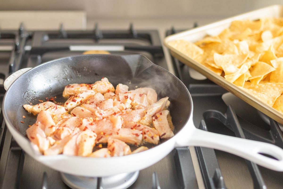 Chicken in a frying pan on a stove, a sheet pan of tortilla chips in the background.