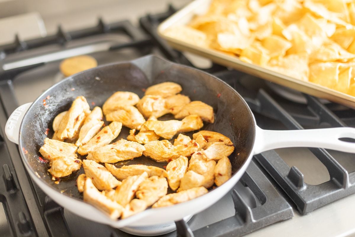 Chicken in a frying pan on a stove, a sheet pan of tortilla chips in the background.