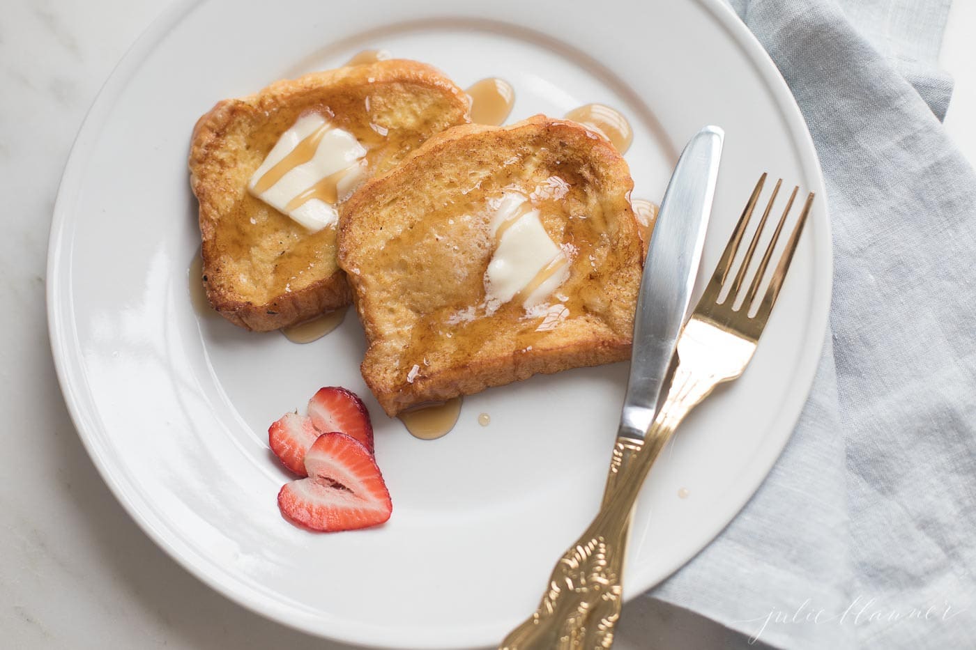 Brioche French toast topped with butter on a white plate, sliced strawberries as garnish.