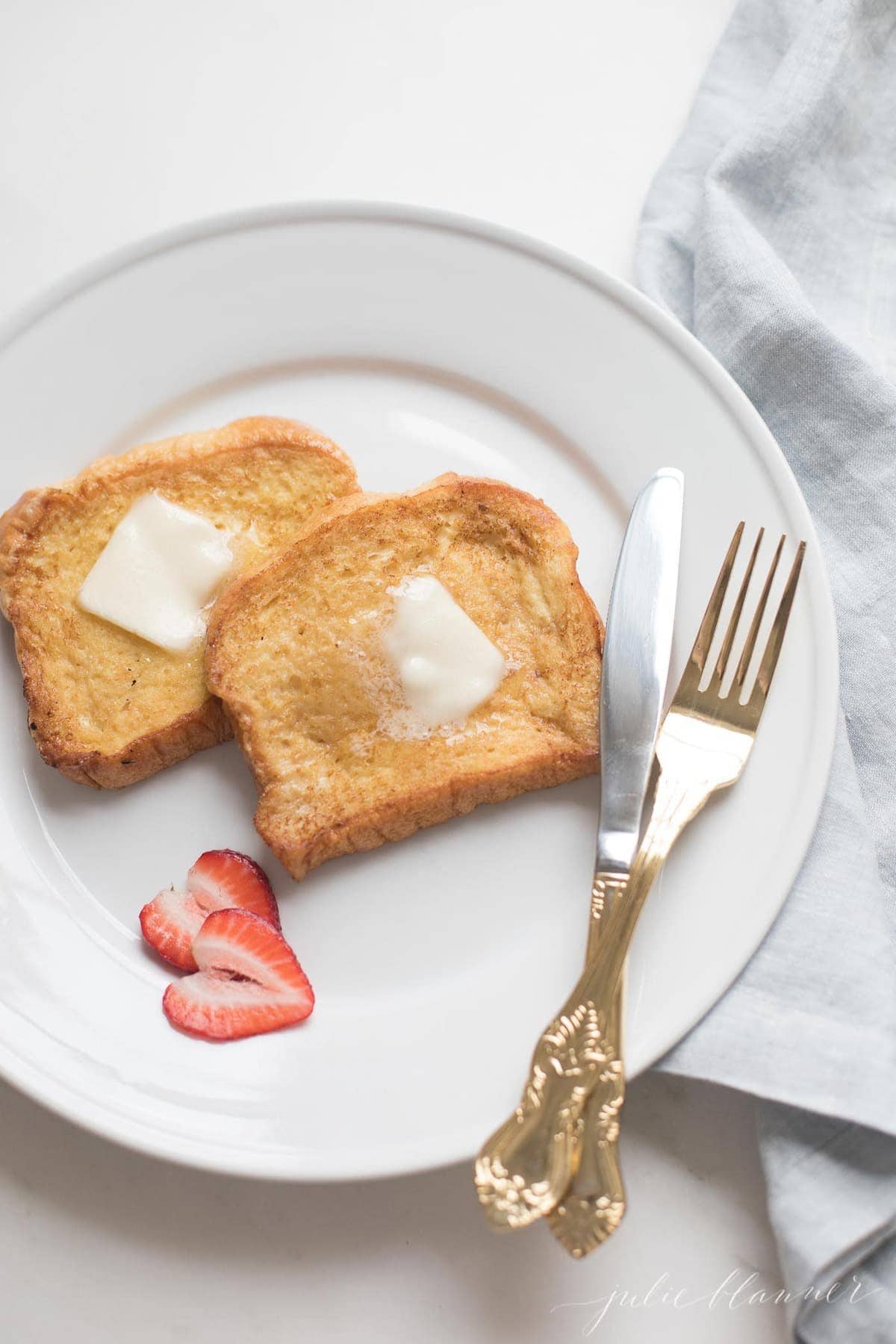 Brioche French toast topped with butter on a white plate, knife and fork to the side.