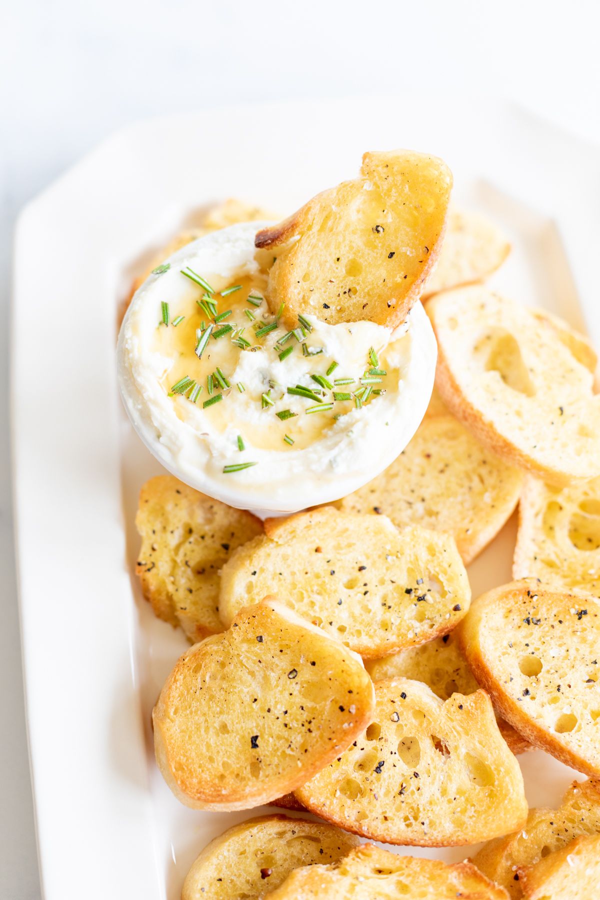 Whipped goat cheese with honey in a white bowl, on a platter of crostini for dipping.