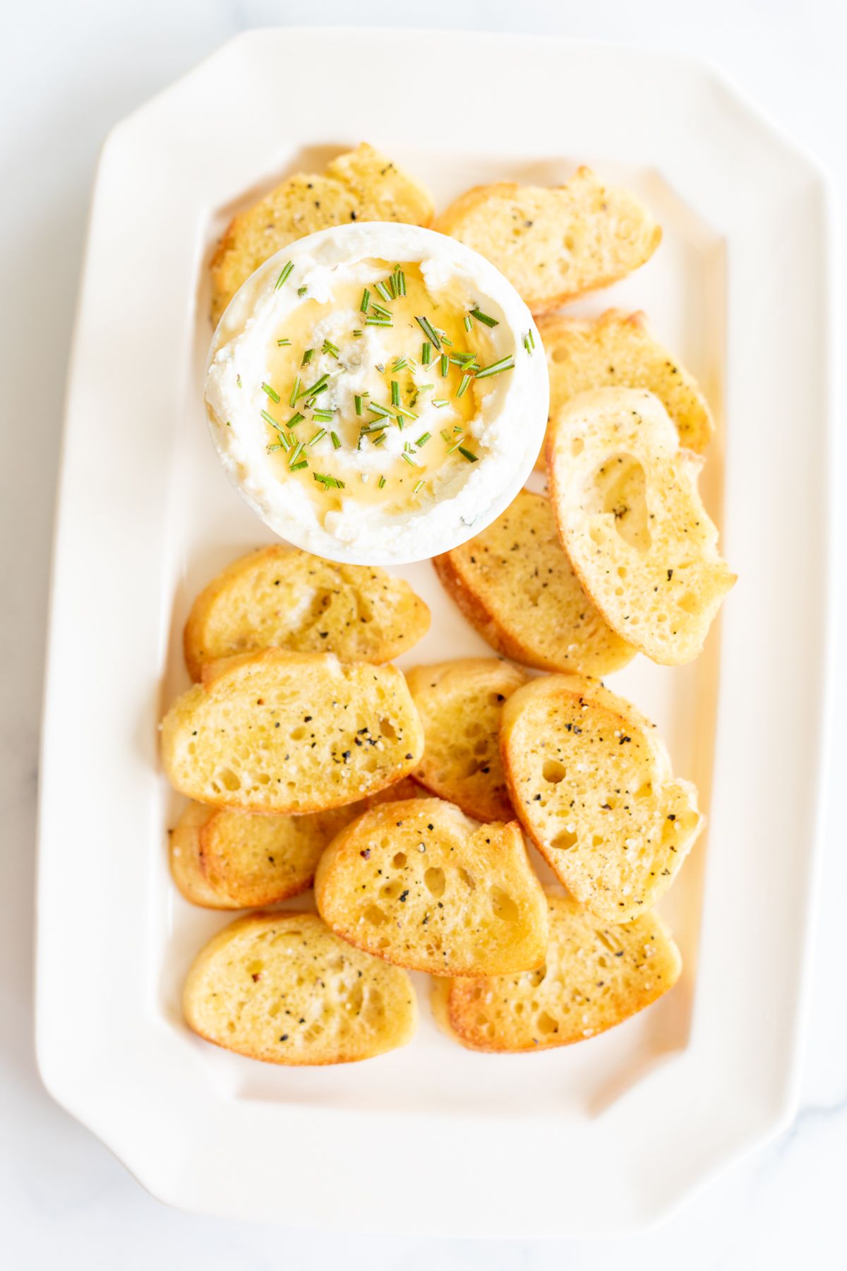 Whipped goat cheese with honey in a white bowl, on a platter of crostini for dipping.