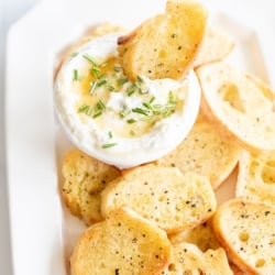 Whipped goat cheese with honey in a white bowl, on a platter of crostini for dipping.