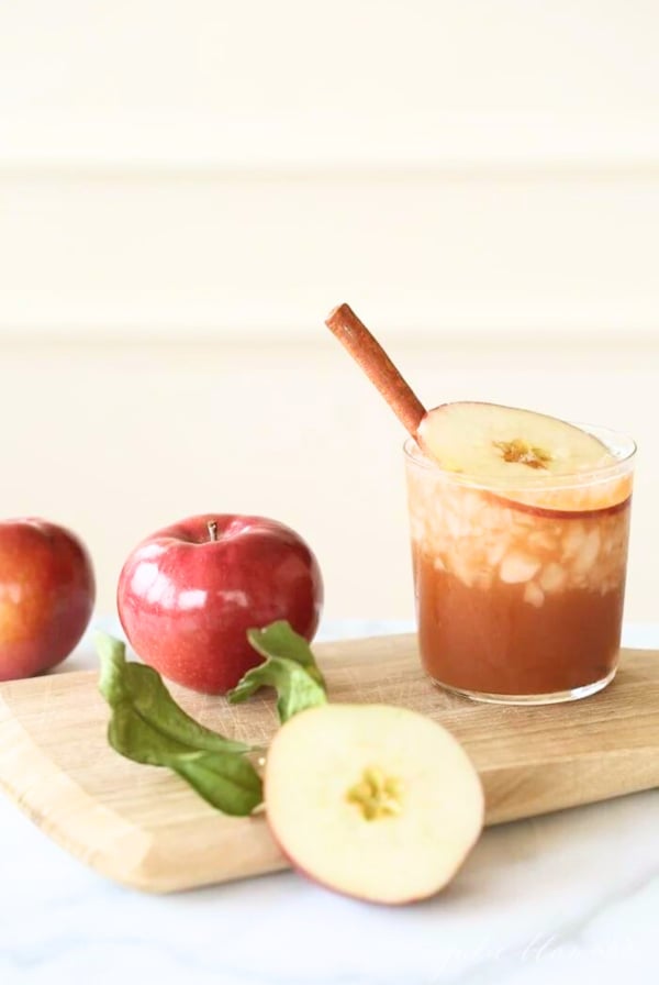 An apple butter old fashioned in a clear glass, apples and a wooden cutting board nearby.