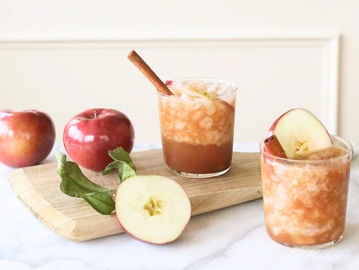 An apple butter old fashioned in a clear glass, apples and a wooden cutting board nearby.