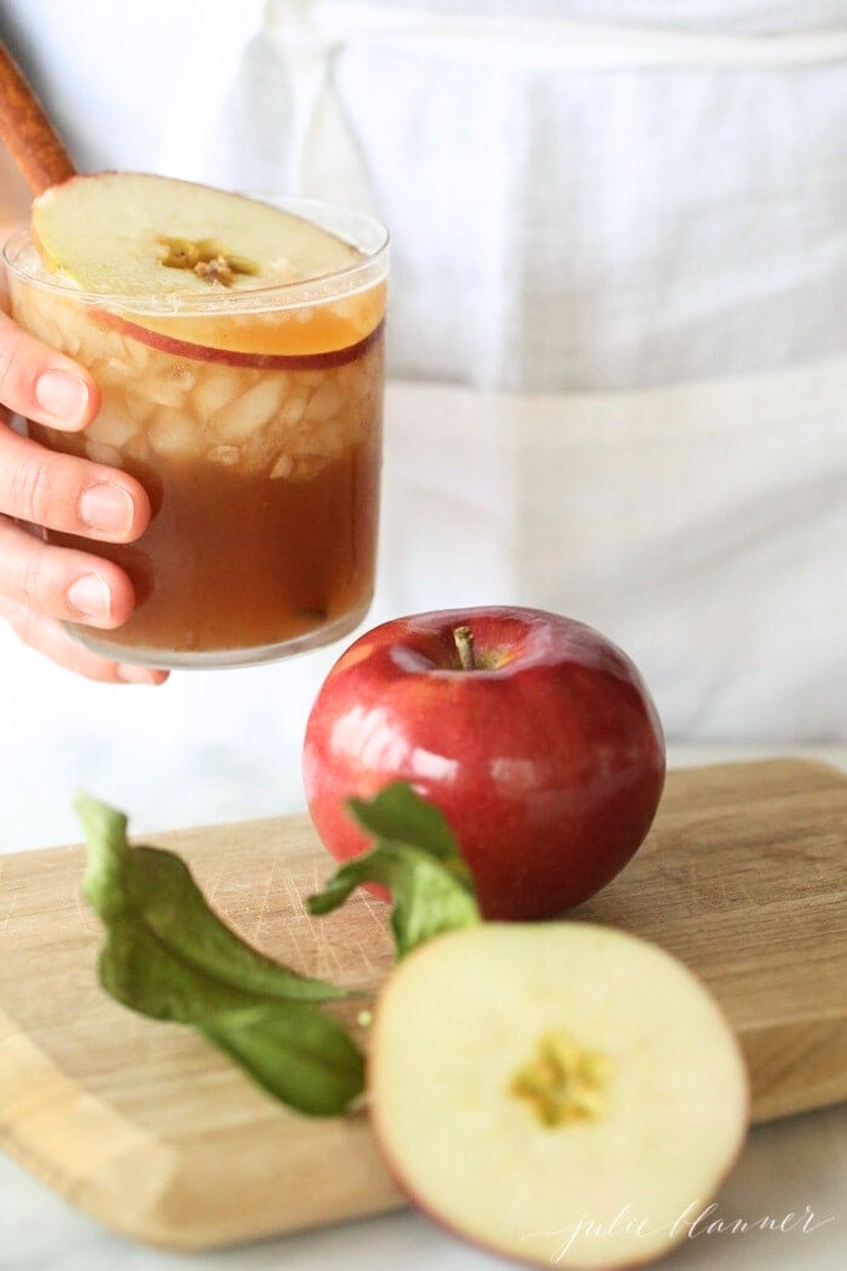 A hand holding an apple butter old fashioned cocktail, with a sliced apple on a wooden cutting board in the foreground