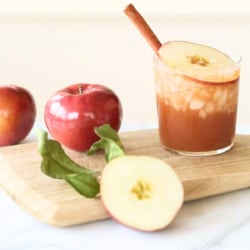 An apple butter old fashioned in a clear glass, apples and a wooden cutting board nearby.