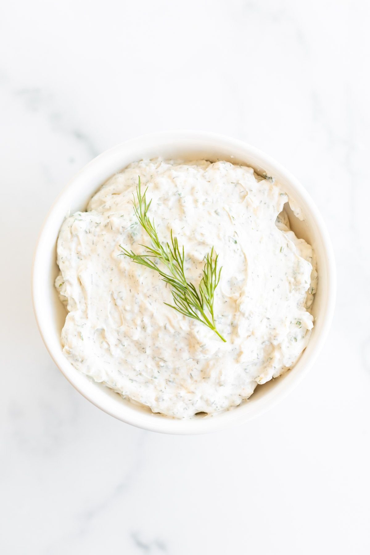 A white bowl on a marble surface, filled with a dill dip recipe and topped with a piece of fresh dill