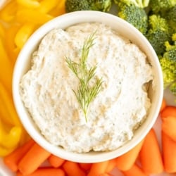 A white bowl full of a dill dip recipe surrounded by vegetables.