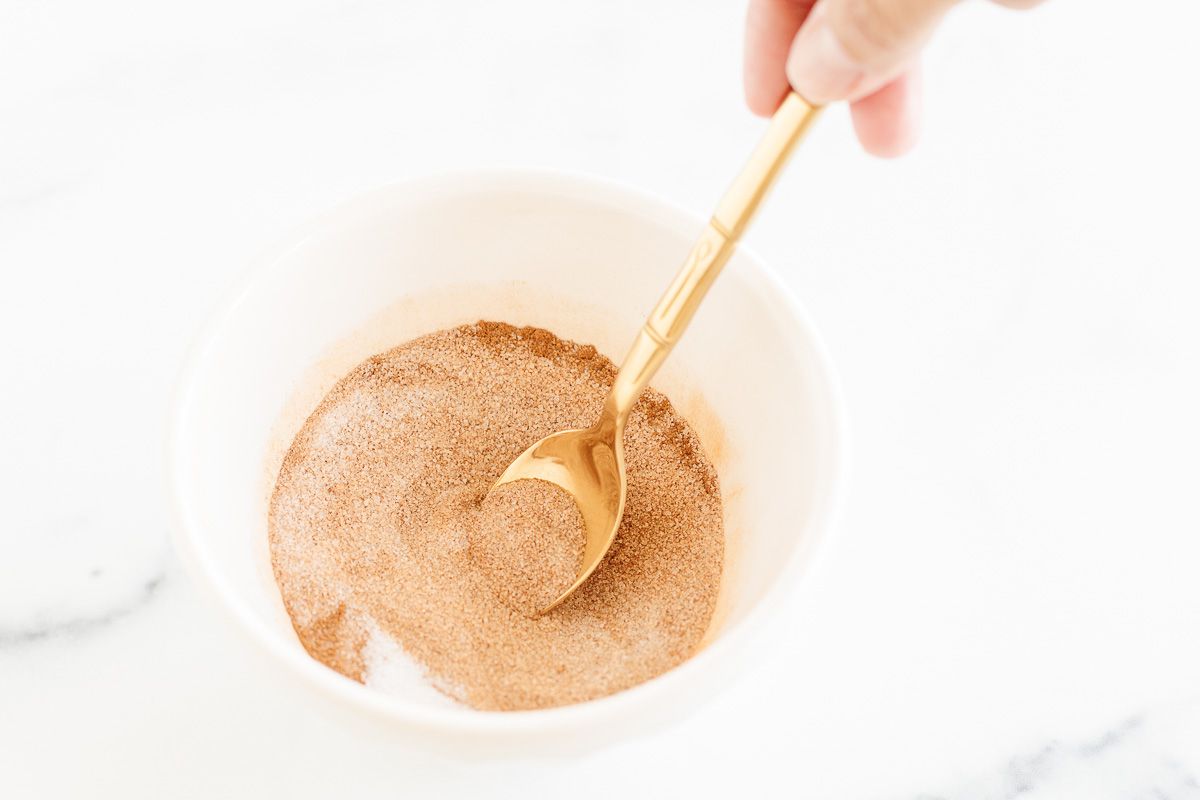 A cinnamon sugar mixture in a white bowl, gold spoon stirring it