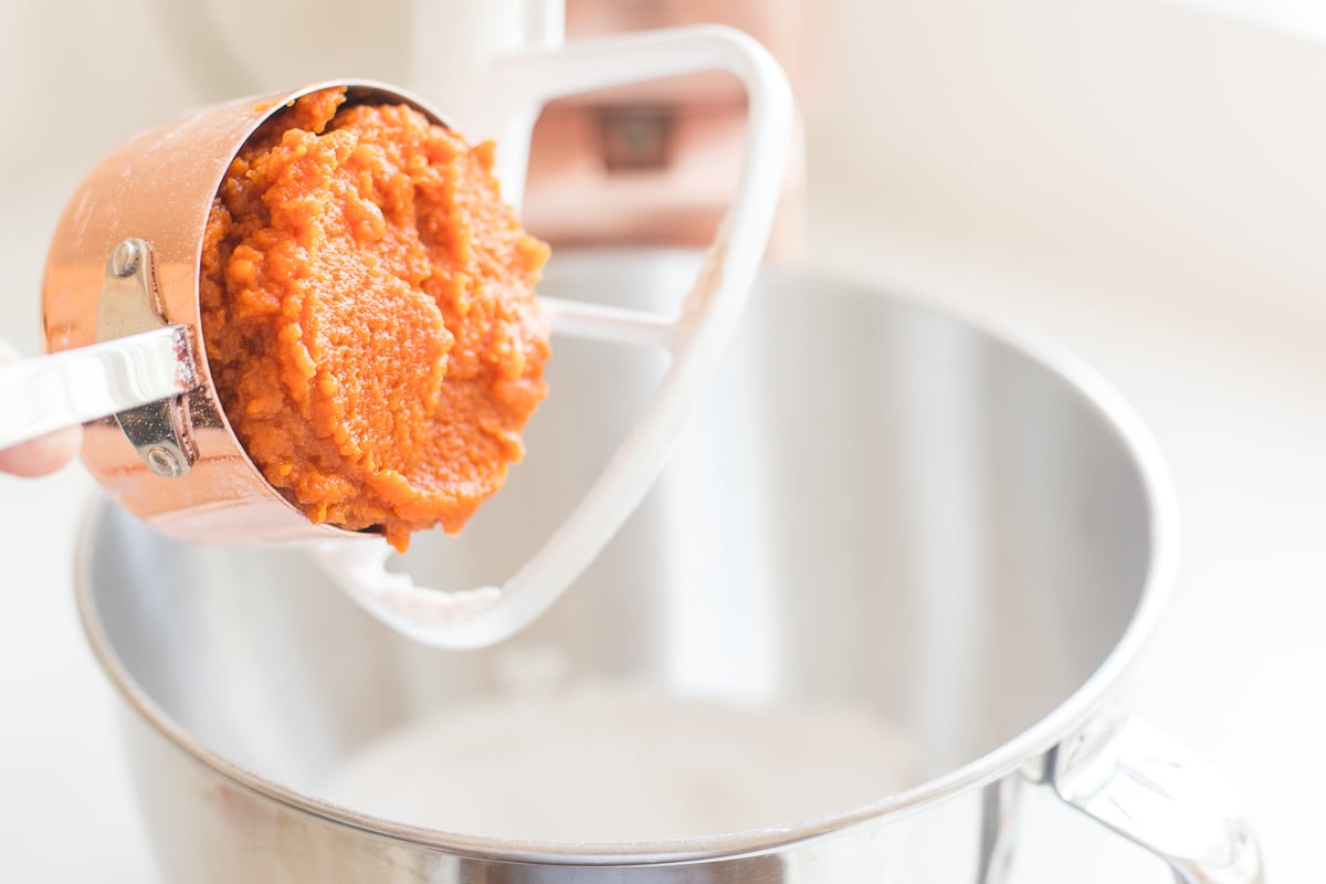 A copper measuring cup adding pumpkin to a stand mixer bowl.