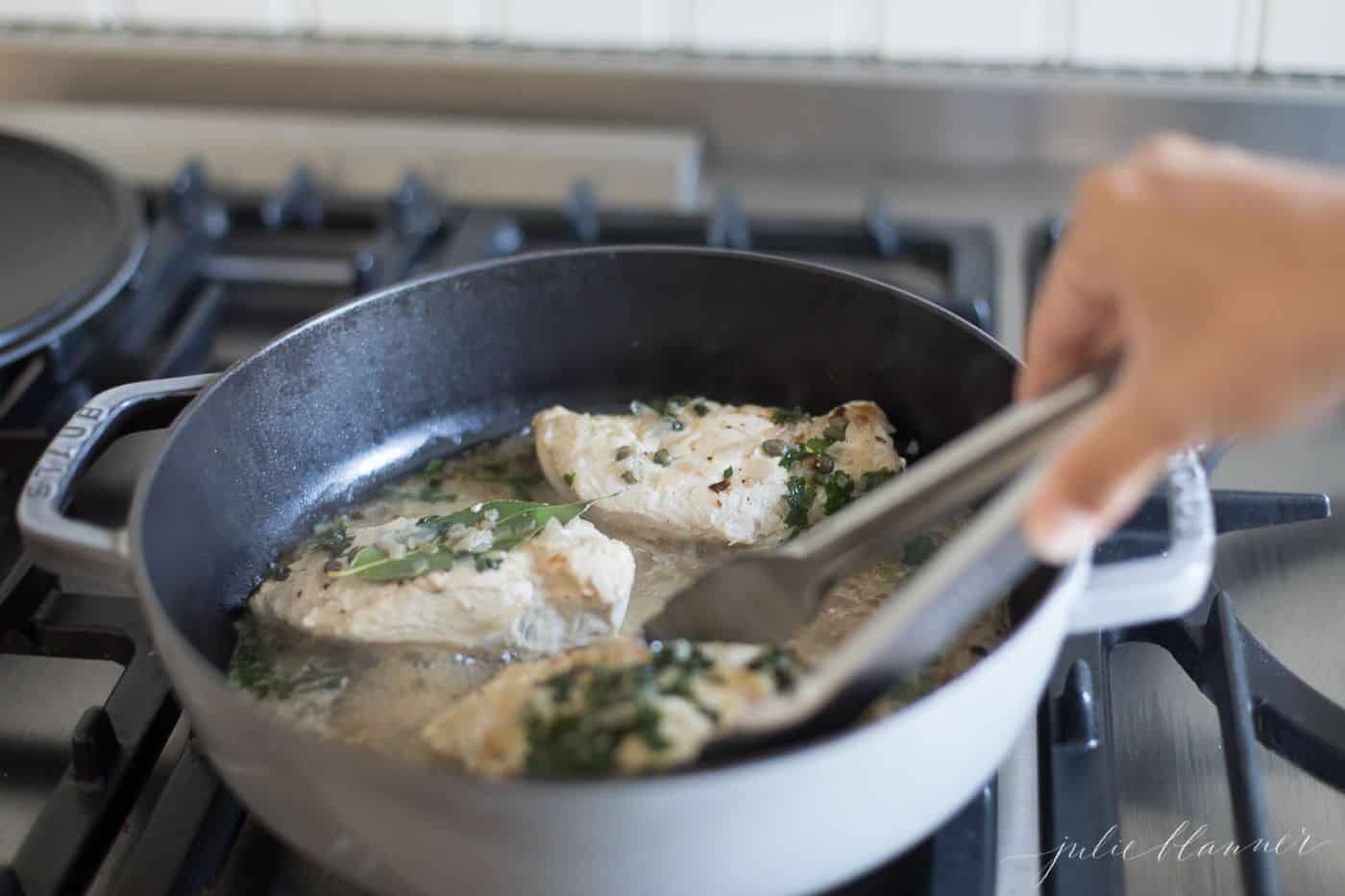 Chicken in white wine sauce in a gray saute pan, hand reaching in with tongs.