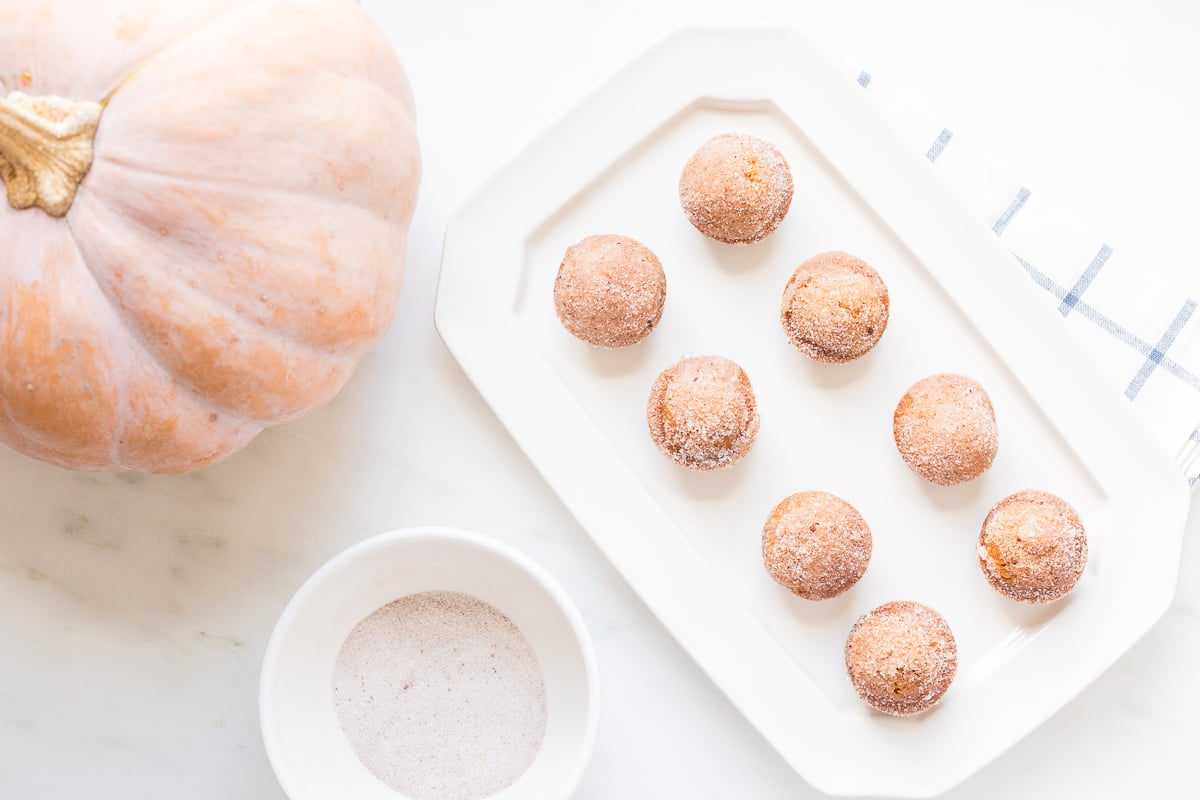 A platter of mini pumpkin muffins with a bowl of sugar and a peach pumpkin in the background. 