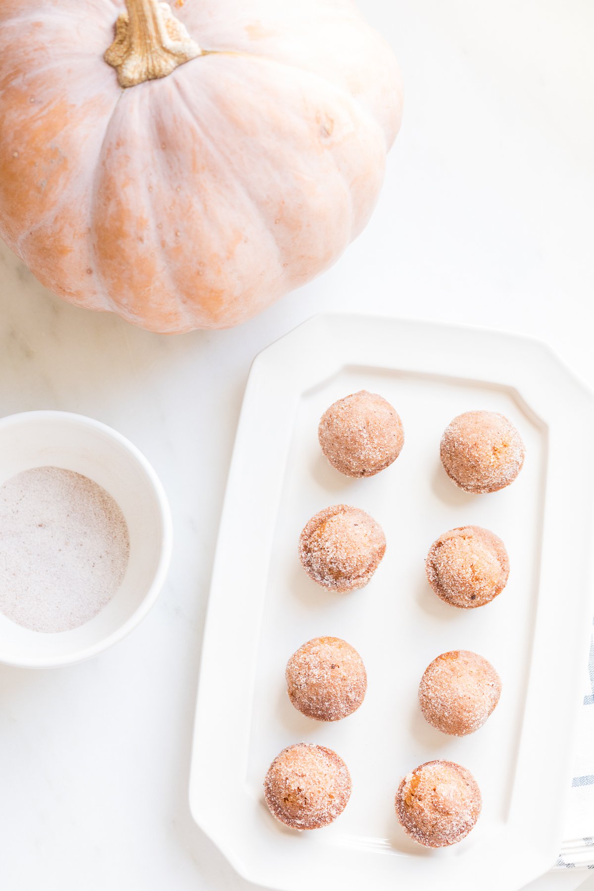 A platter of mini pumpkin muffins with a bowl of sugar and a peach pumpkin in the background.