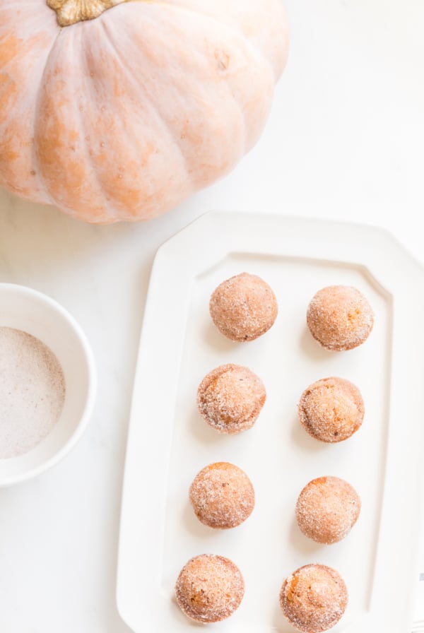 A platter of mini pumpkin muffins with a bowl of sugar and a peach pumpkin in the background.