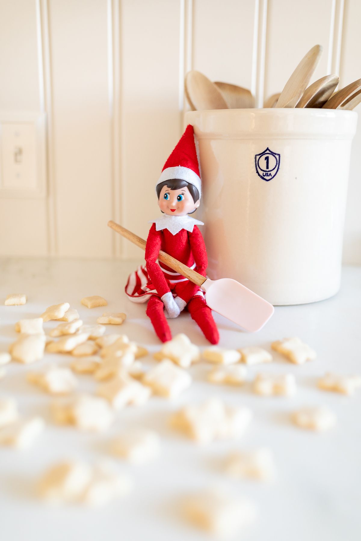 An elf on the shelf sitting on a kitchen counter, with tiny shortbread elf on the shelf cookies surrounding her.