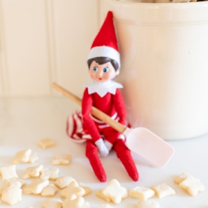 An elf on the shelf sitting on a kitchen counter, with tiny shortbread elf on the shelf cookies surrounding her.