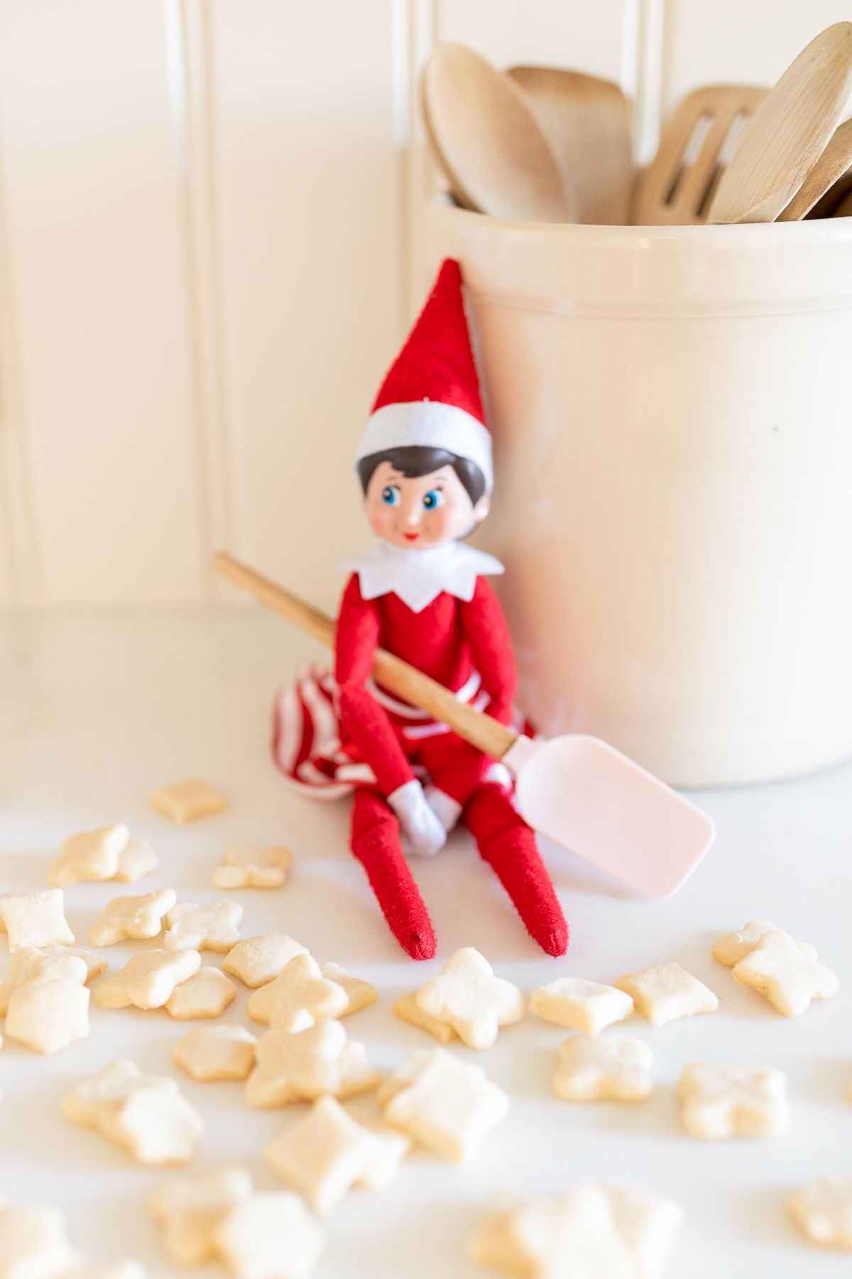 An elf on the shelf sitting on a kitchen counter, with tiny shortbread elf on the shelf cookies surrounding her. 