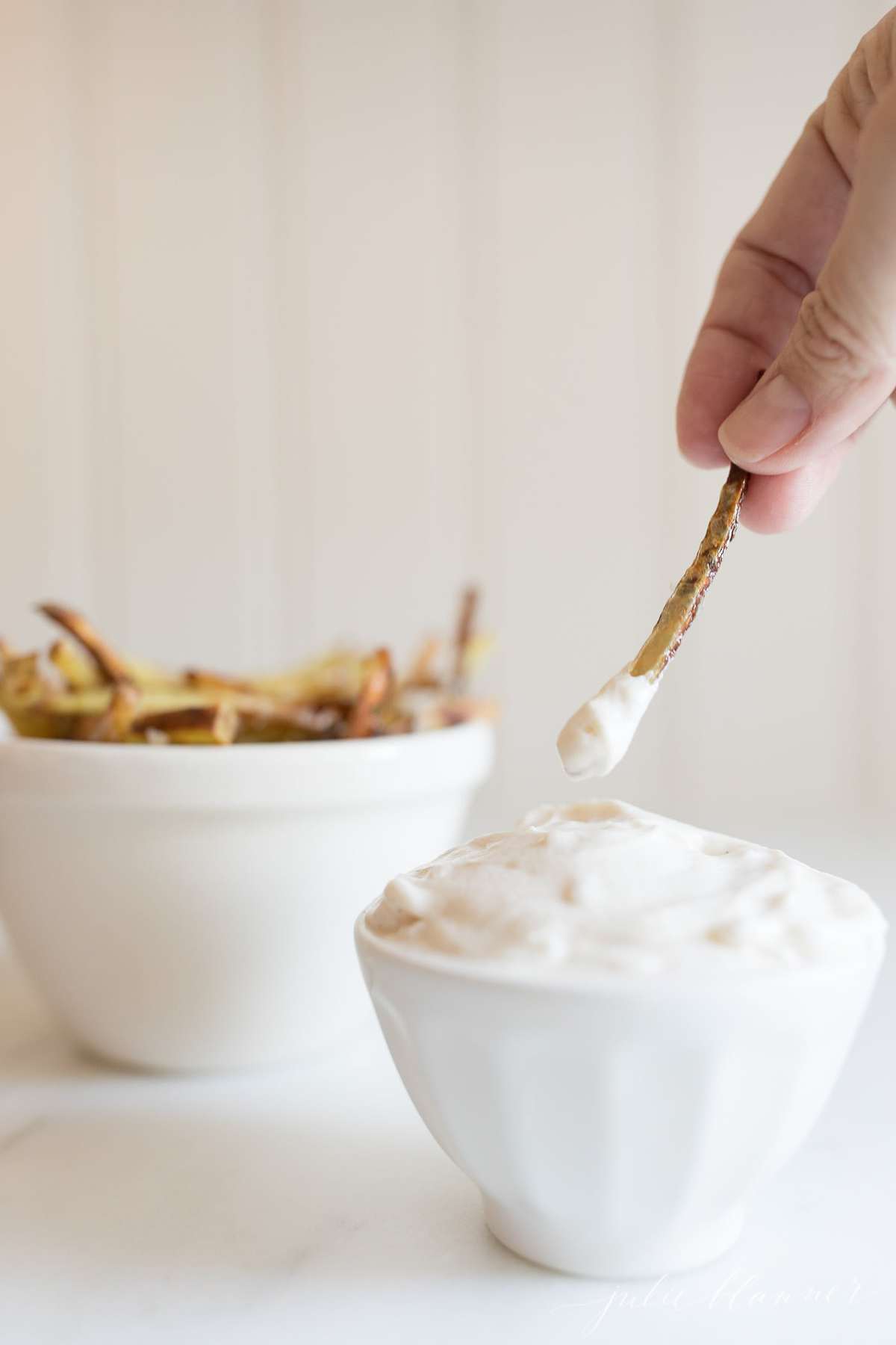 truffle mayo in a white bowl with a bowl of fries in the background, hand reaching in to dip with a single fry