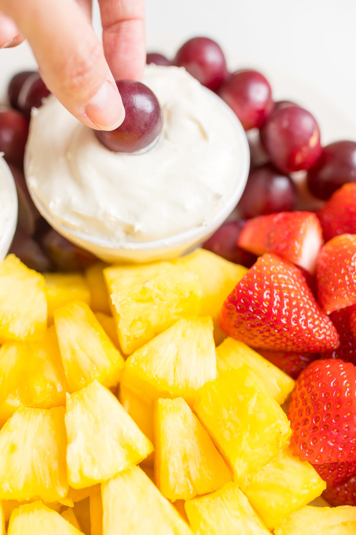 A hand dipping a grape into a bowl of fruit dip on a fruit platter