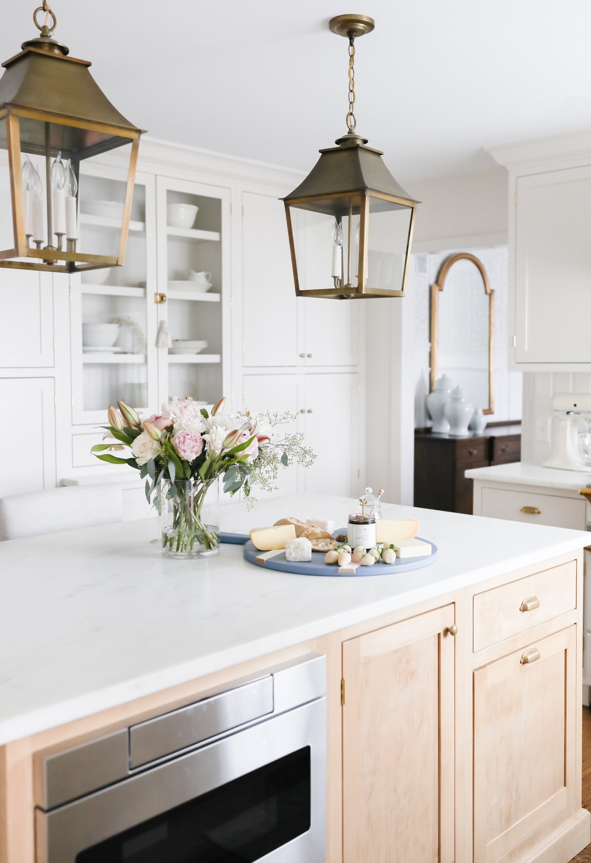 A modern kitchen showcases a white marble island adorned with a vase of flowers and a cheese platter. Two brass pendant lights hang overhead, while well-planned kitchen organization is evident in the white cabinets providing ample storage in the background.