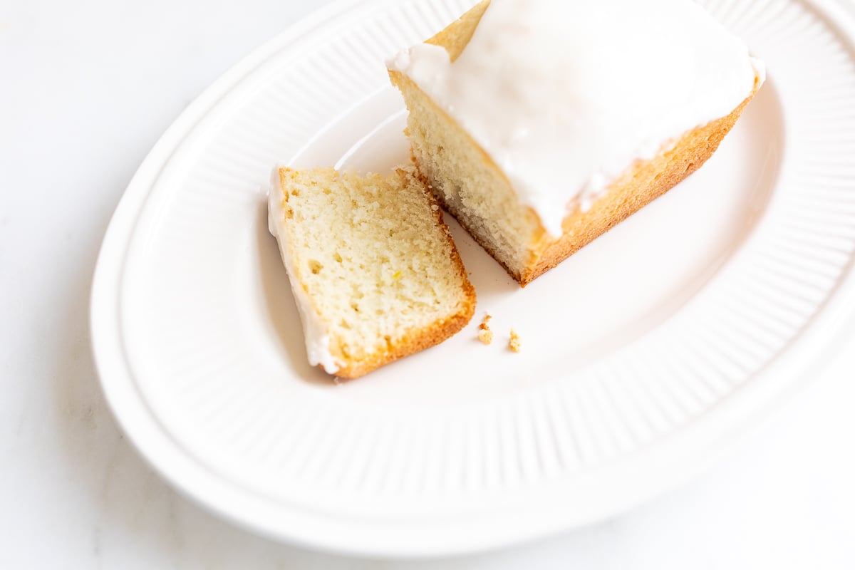 A frosted lemon loaf on a white platter, one slice cut.