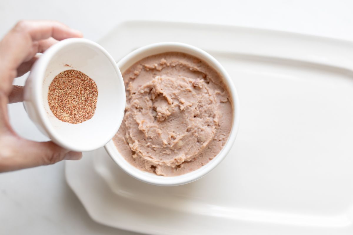 A hand mixing taco seasoning into a white bowl of refried beans.