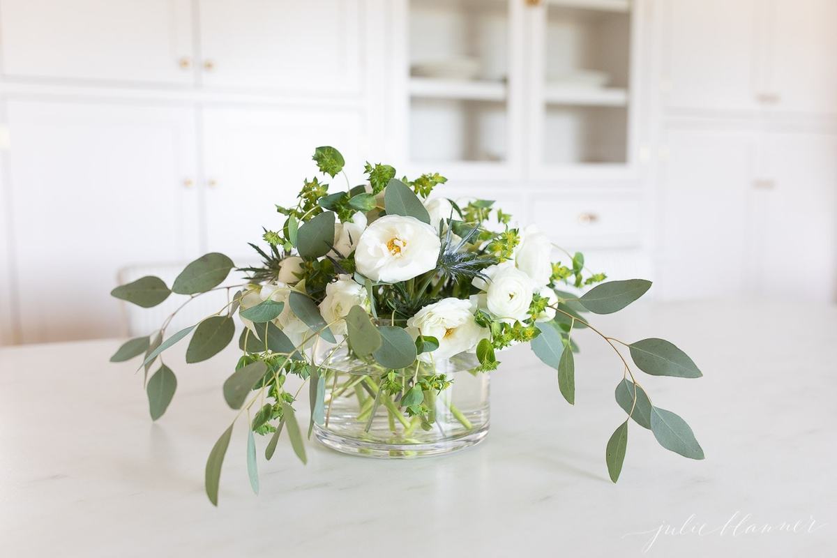 A bouquet of white ranunculus in a glass vase on a white kitchen countertop. 