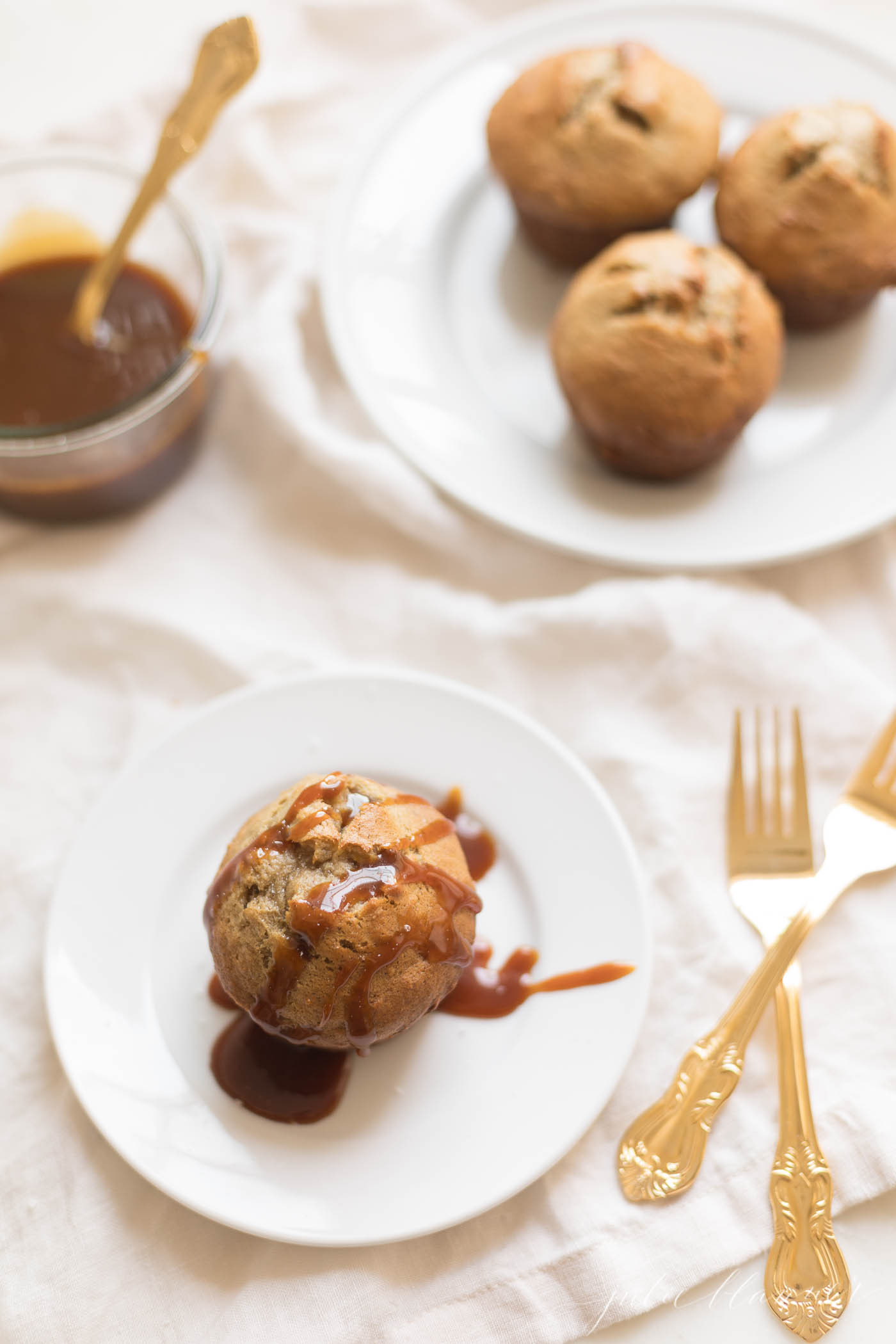 Caramel and Banana Muffin topped with caramel on a white plate, gold silverware to the side.